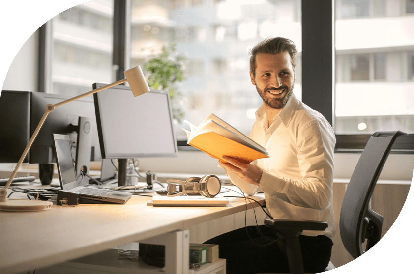 Man sat at a desk smiling holding an orange notebook.